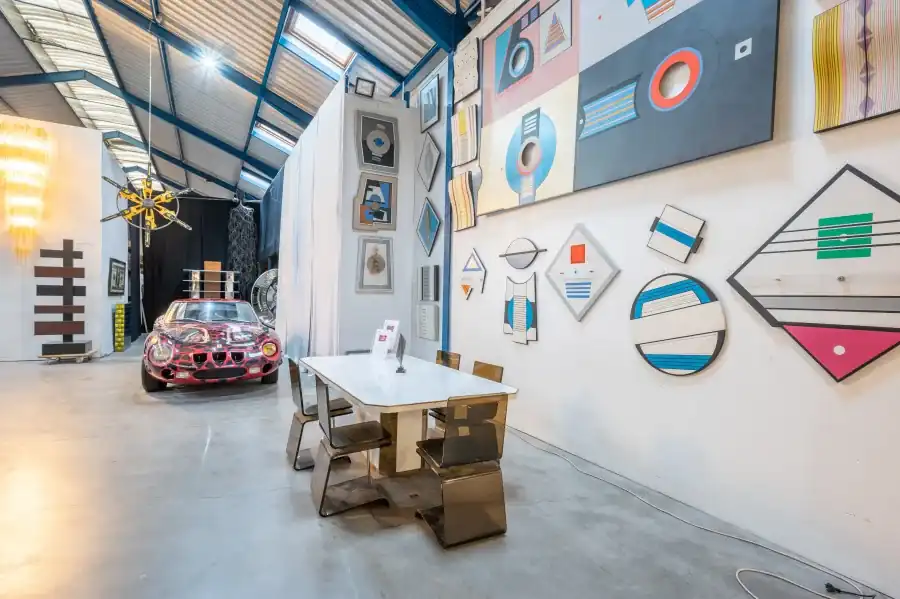 Halle sous charpente métallique, table et chaises devant un mur d’œuvres géométriques